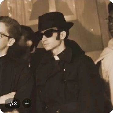 Man in dark clothing and hat with sunglasses at a formal event.