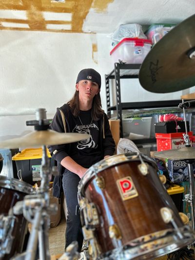 Young man sits behind a drum set in a cluttered garage.