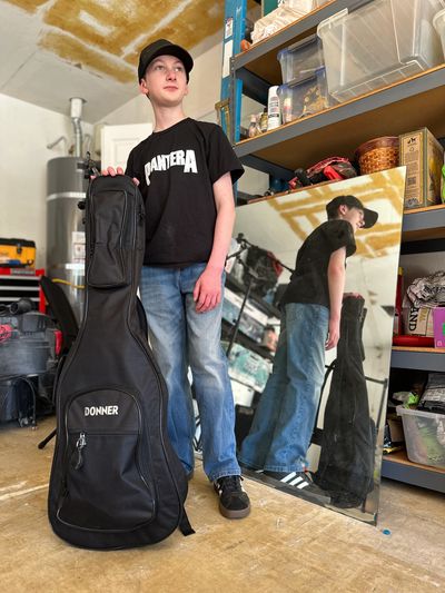 Teen boy holding a guitar case, standing next to a large mirror in a garage.