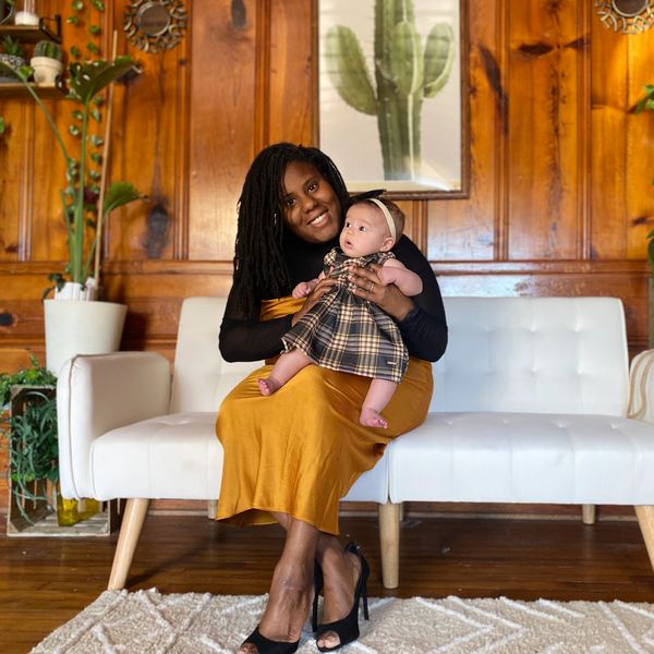 A woman in a mustard dress holds a baby in a plaid dress on a white couch.