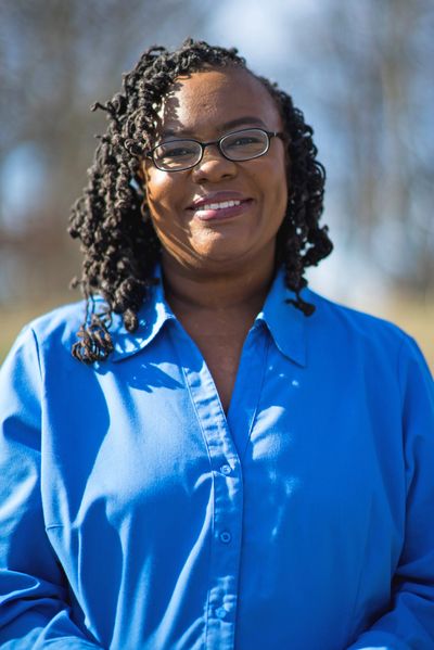 A smiling woman with glasses wearing a blue shirt stands outdoors in sunlight.