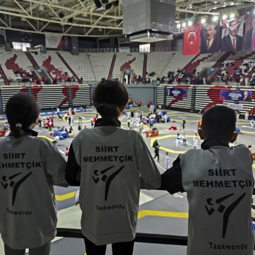 Siirtli üç taekwondo sporcusu büyük salonda yarışmayı izliyor.