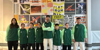 A group of eight martial artists in green uniforms stand in front of a sports-themed wall.