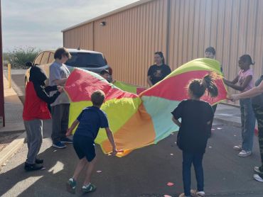 Children playing with a colorful parachute outside near a building.