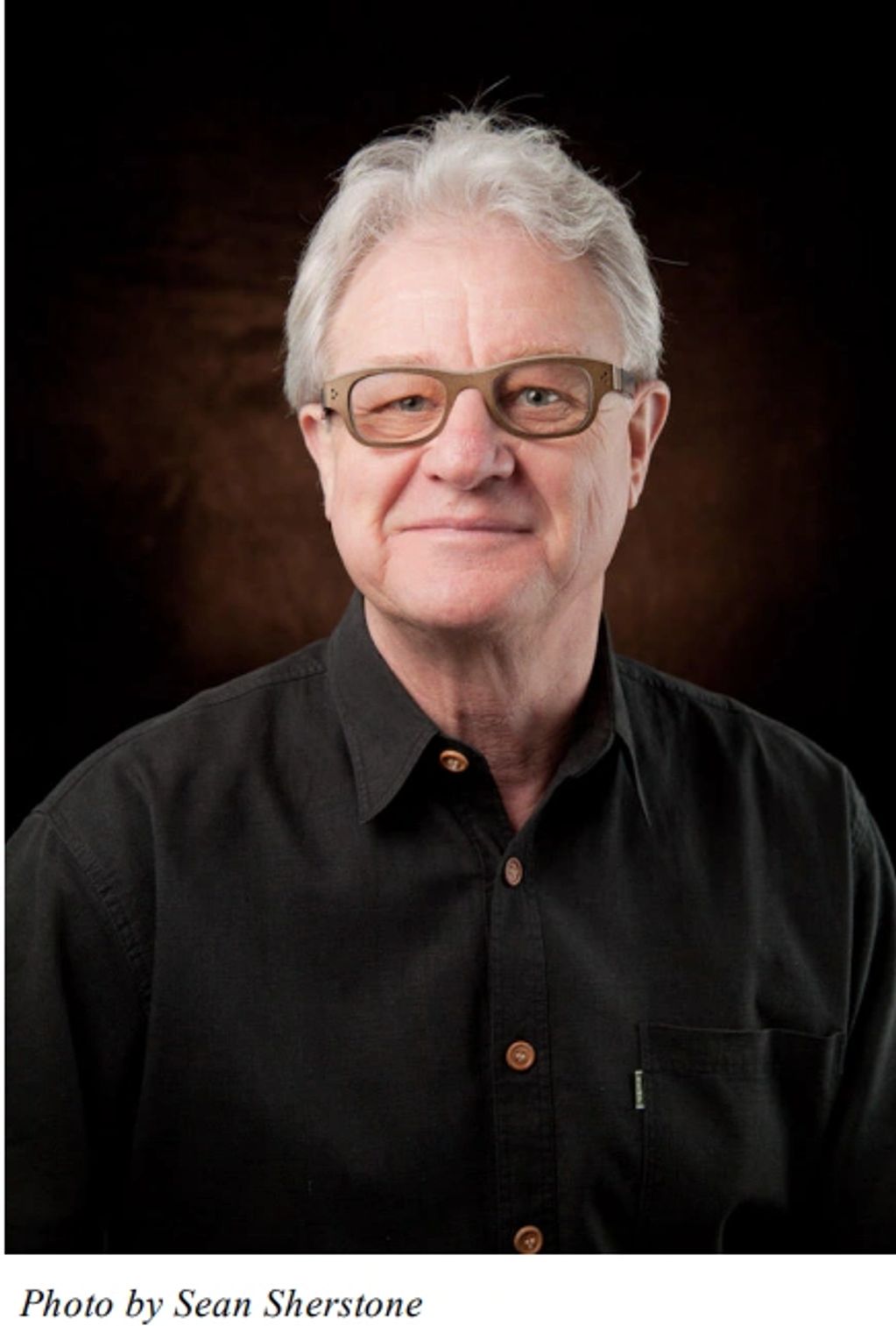 Portrait of an older man with white hair and glasses wearing a black shirt.