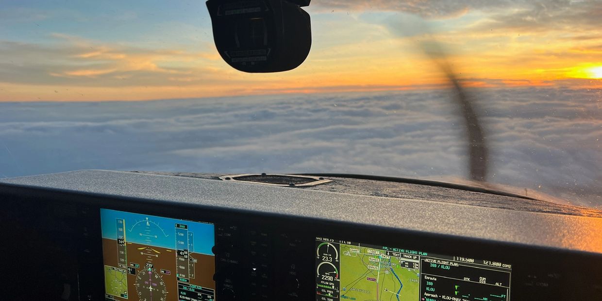 View from inside a small aircraft cockpit flying above the clouds during sunset.