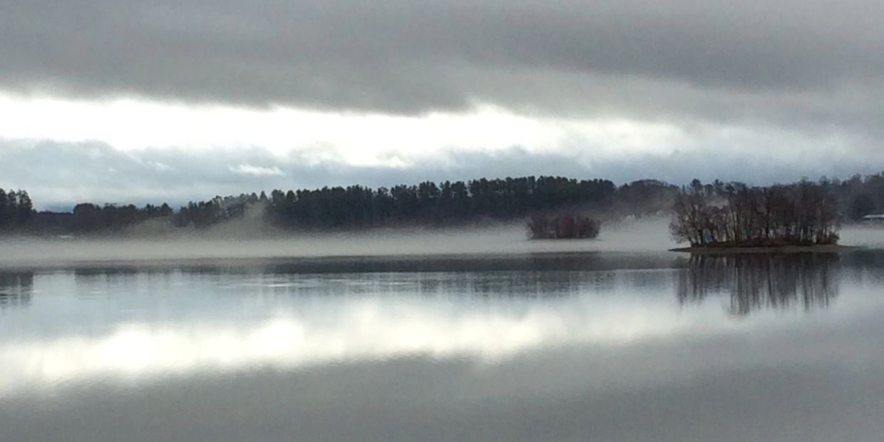 Foggy lake with small tree-covered islands under cloudy sky.