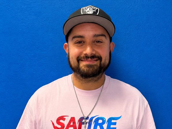 Smiling man wearing a Raiders cap and graphic T-shirt against a blue wall.