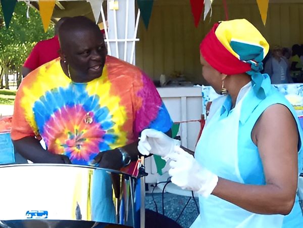 Stanley Alleyne wearing a colorful tie-dye shirt plays a steel drum, while a woman with gloved hands