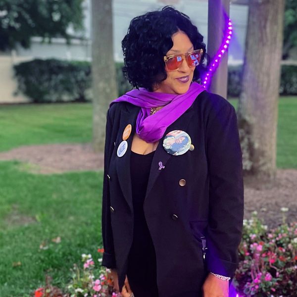 Woman wearing a black coat, purple scarf, and sunglasses outdoors.
