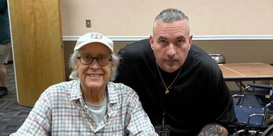 Two men posing together indoors, one wearing a cap and glasses.
