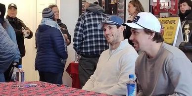 Two men smiling and sitting at a table in a busy indoor setting.