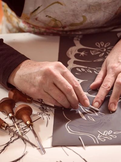 Close-up of hands carving a linoleum printmaking block with a wood-handled gouge, surrounded by curl