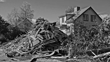 A large pile of wooden debris in front of a house with trees in the background.