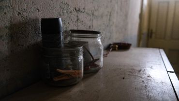 Dimly lit room with jars and a metal cup on a wooden surface near a textured wall.