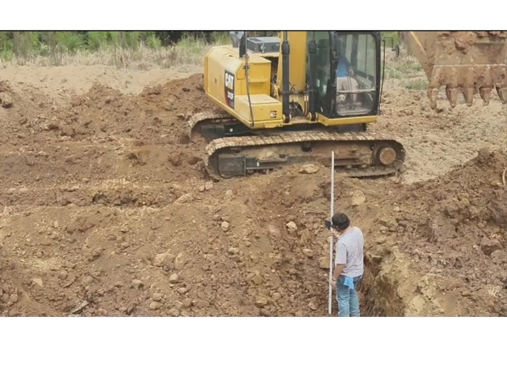 guy standing in a hole measuring depth while a CAT excavates beside him