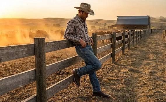 Cowboy leaning against a fence at sunset on a ranch.