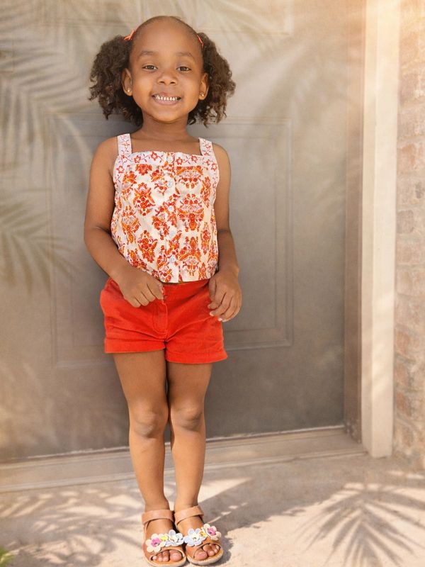 Smiling girl in floral top and red shorts stands outside on sunny day.