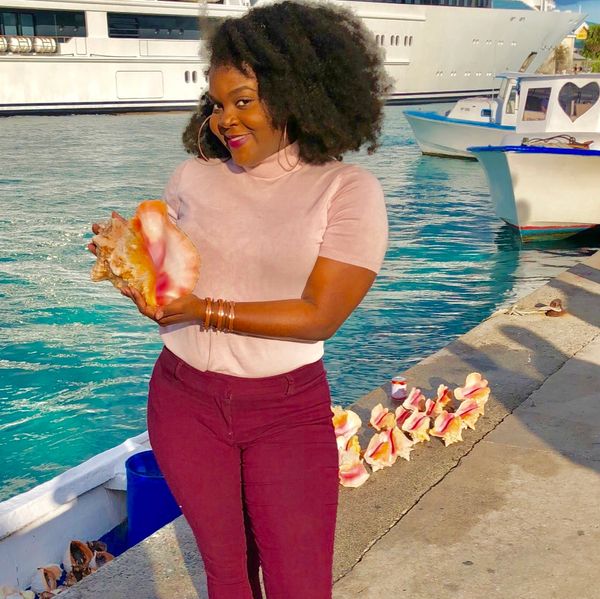 Woman holding a large seashell by the waterfront with boats in the background.