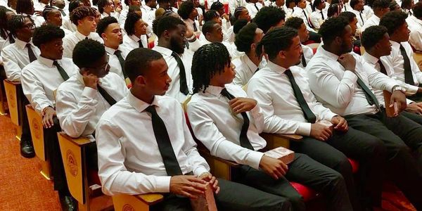 Young men in white shirts and black ties seated in an auditorium.