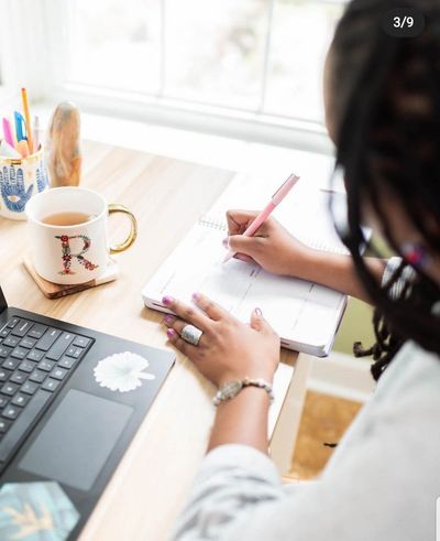 Person writing in a planner at a desk with a laptop and coffee mug.