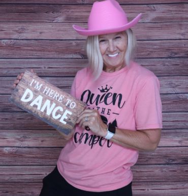Smiling woman in pink hat and shirt holds a sign saying 'I'm here to dance'.