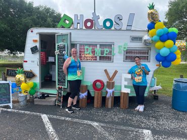 Two women stand in front of a decorated photo booth trailer with colorful balloons and large letters spelling PHOTO and LOVE.