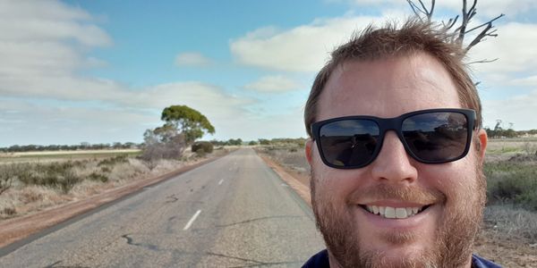 Smiling man with sunglasses on a deserted rural road under a blue sky.