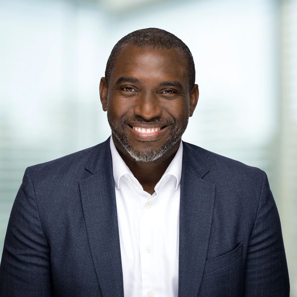 Smiling man in a navy suit and white shirt against a blurred background.