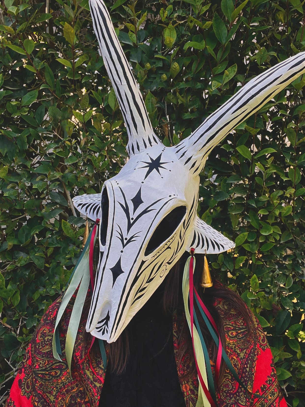 Person wearing a large white horned mask with black designs and colorful ribbons.