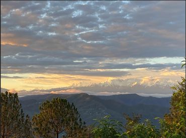 Sunlit mountain range under a cloudy sky with lush trees in the foreground.