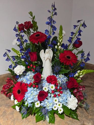 Vibrant floral arrangement with red, blue, white flowers and an angel centerpiece.