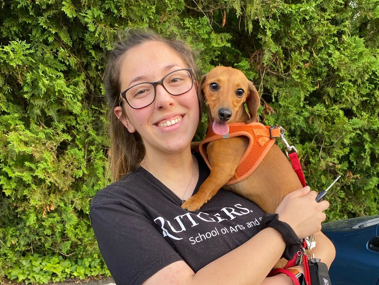 Young woman smiling and holding a small dog in an orange harness outdoors.