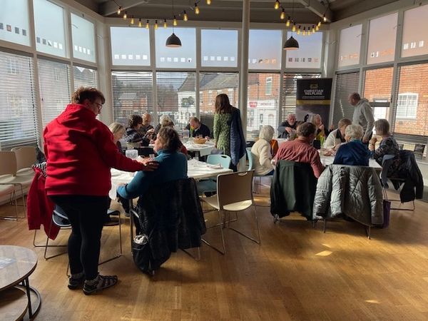 Group of elderly people socializing around tables in a sunlit room.