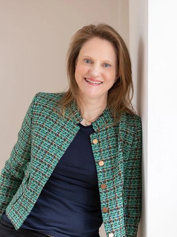 Smiling woman in a green textured jacket leaning against a white wall.