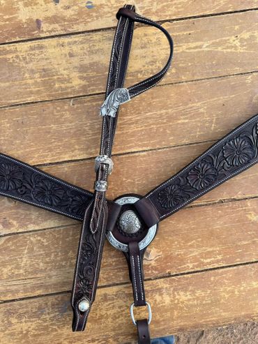 Intricately tooled dark leather horse breast collar with silver accents on wooden surface.