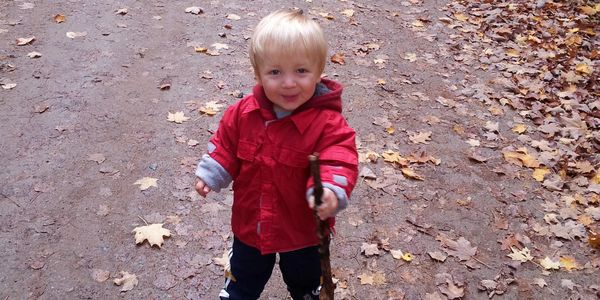 Young child in a red jacket holding a stick on a leaf-covered path.