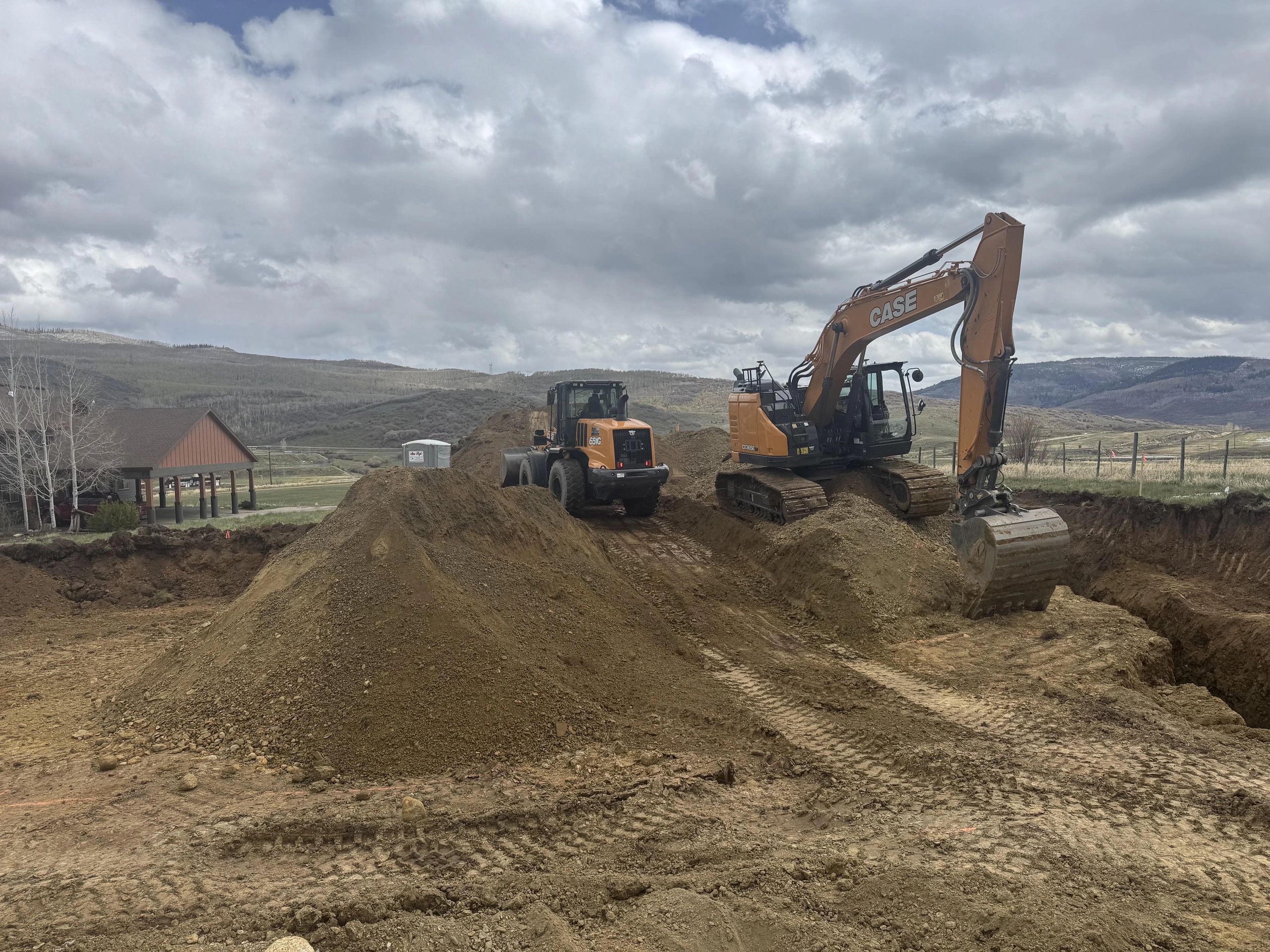 Excavator and loader working on a dirt construction site with cloudy skies.