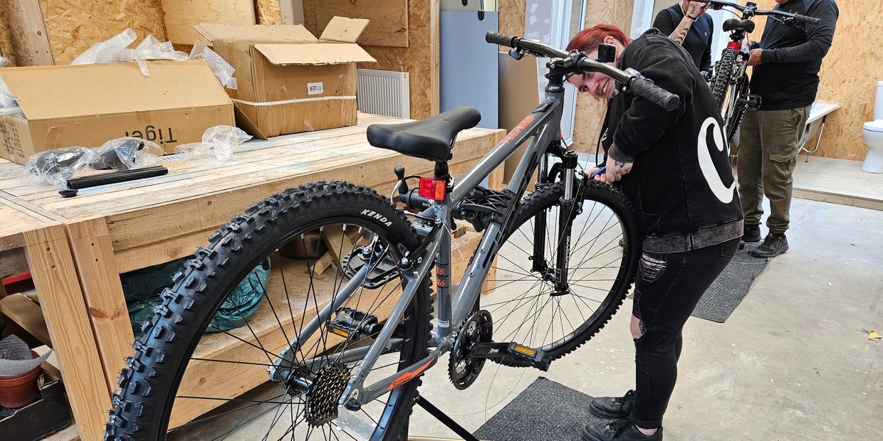 A person fixing a mountain bike in a workshop with another person nearby.