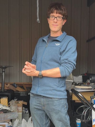 Man in a blue jacket and glasses standing in a cluttered garage.