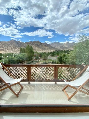 Two wooden lounge chairs on a balcony overlooking mountains under a partly cloudy sky.