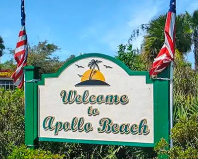 Welcome sign for Apollo Beach with palm trees and American flags.