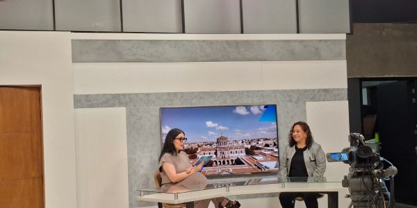 Two women engaged in a TV interview in a studio with a cityscape backdrop.