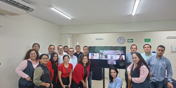 Group of professionals posing in an office with a video conference screen.