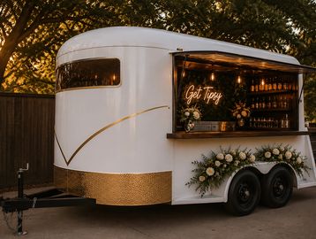 A white and gold mobile bar trailer decorated with flowers and a neon sign.