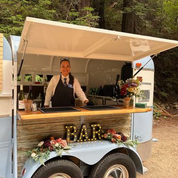 Smiling bartender at outdoor mobile bar with floral decorations and cocktail menu.