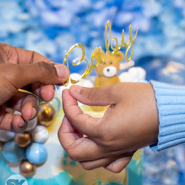 Hands holding a golden 'Baby' decoration with a teddy bear and balloons in the background.