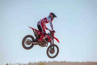 A motocross rider mid-air on a red dirt bike against a clear sky.