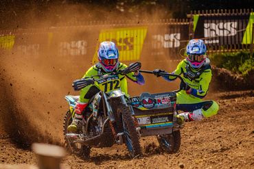 Two motocross racers in neon gear speed through a dirt track, kicking up dust.