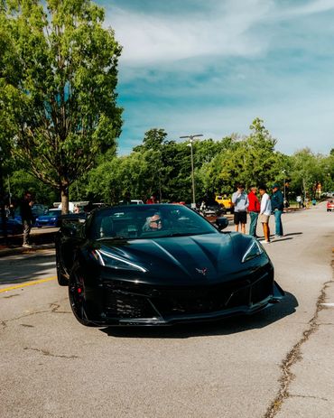 Black sports car driving on a sunny street with people nearby.